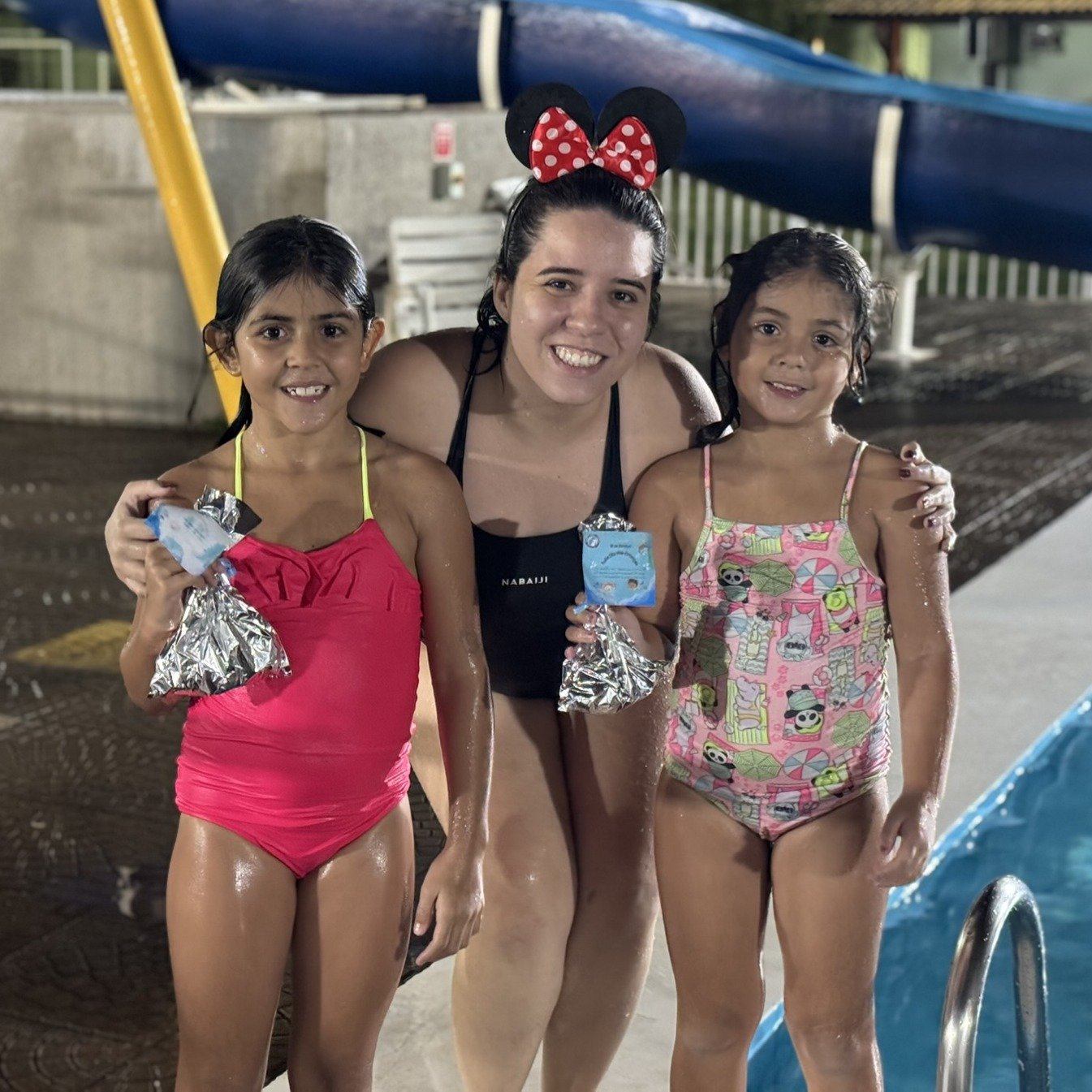 2 alunas de natação fazendo pose para a foto juntas a professora Isabella, mostrando a lembrancinha que ganharam no dia das crianças