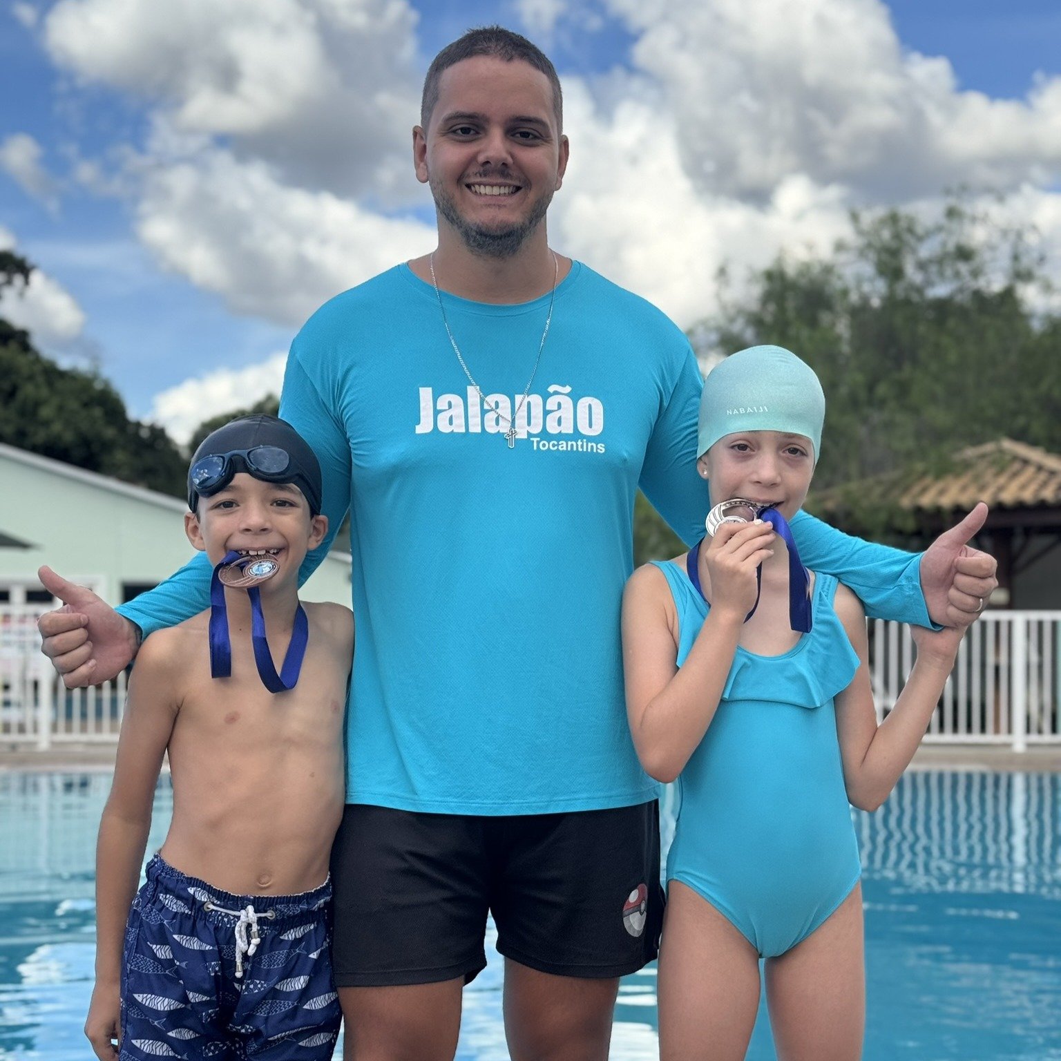 2 alunos de natação fazendo pose para a foto juntos ao professor Filipe, mostrando as medalhas que ganharam no rankeamento interno da natação